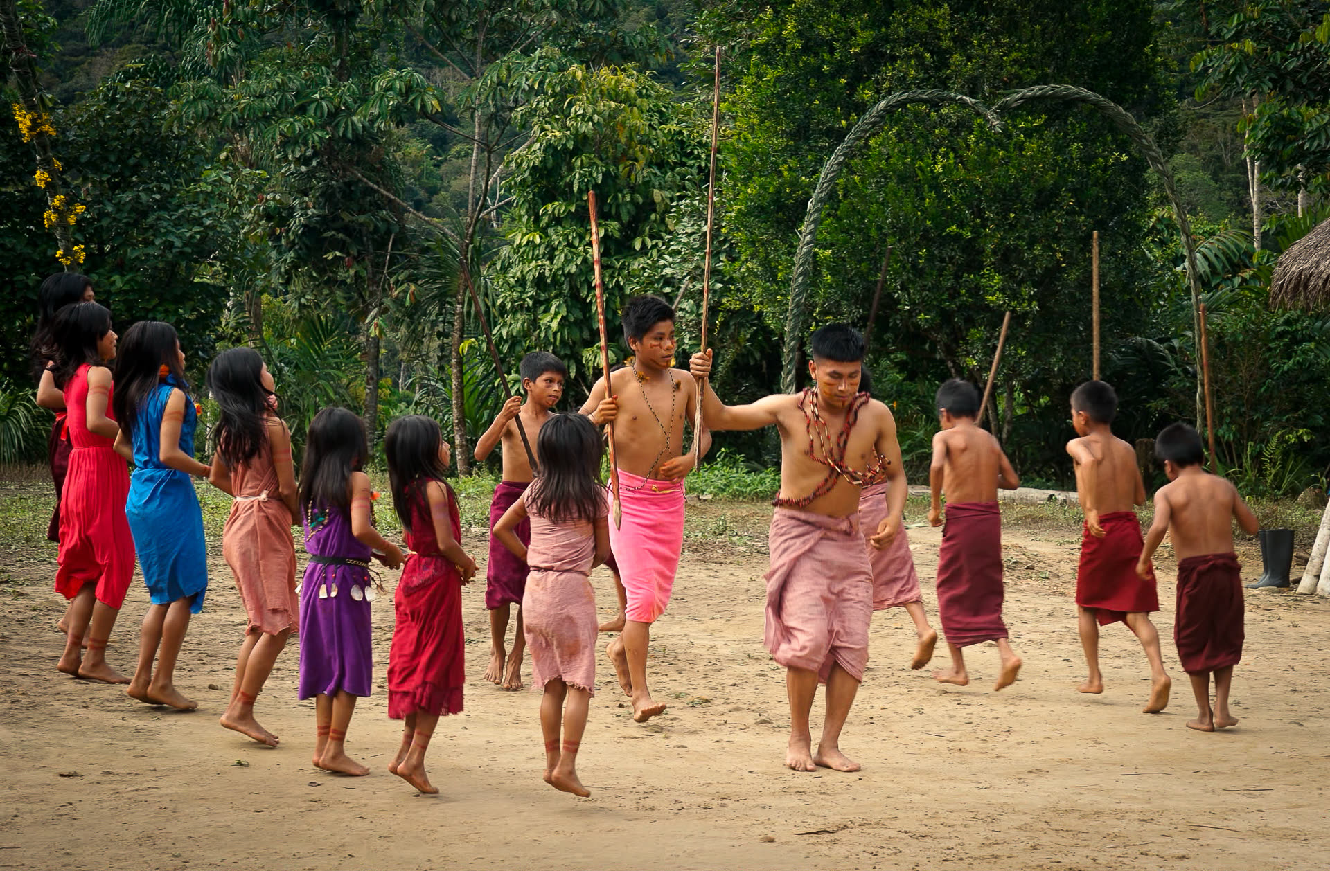 Indigenous children and young men performing a traditional dance barefoot on sandy ground, rainforest behind them
