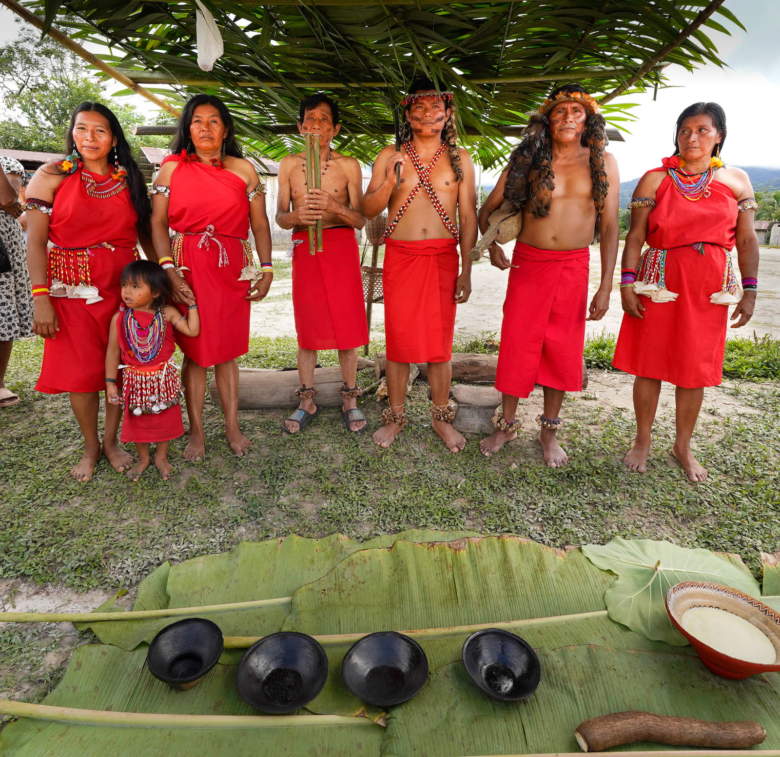 Awajún women, men and a child in traditional dress beside a palm-leaf shelter